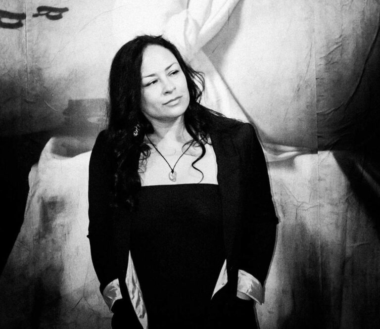 A black-and-white photograph of Tracy Rector, a woman with long black hair, with her hands in her pockets.