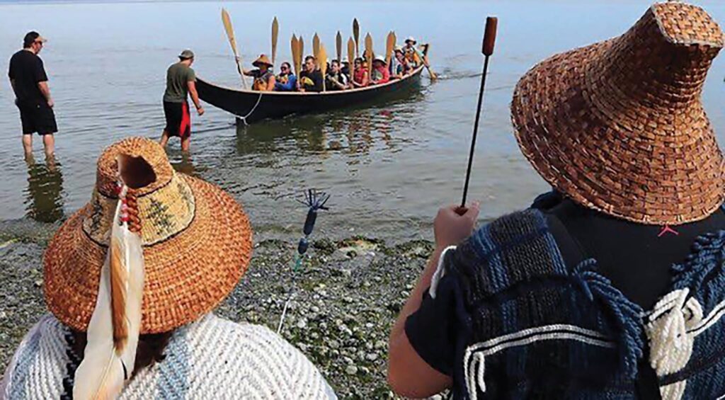 Two individuals in woven hats stand with their backs to the camera looking towards a body of water where a boat full of rowers is coming towards the shore.