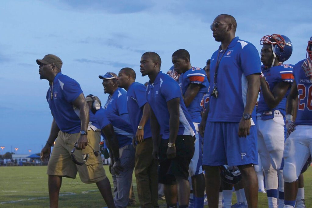 Football team watching the game from the sidelines. They wear royal blue jerseys and are leaning in to shout to the team. All of the visible coaches and players have dark skin.