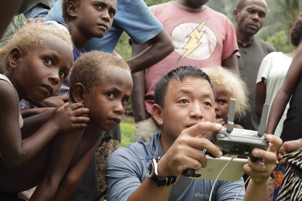A man with medium light skin and black hair holds a remote control and is looking past the camera. He is surrounded by children and adults with dark skin and light brown or blonde hair who are all also looking past the camera at what the other man is controling.