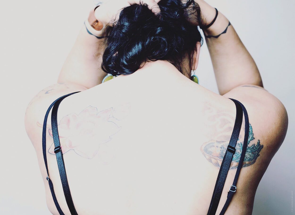 The back of a woman who is facing away from the camera with her hands on top of her head. She has light skin, black hair and tattoos on her back and arms. She has 4 black straps coming from her clothes.