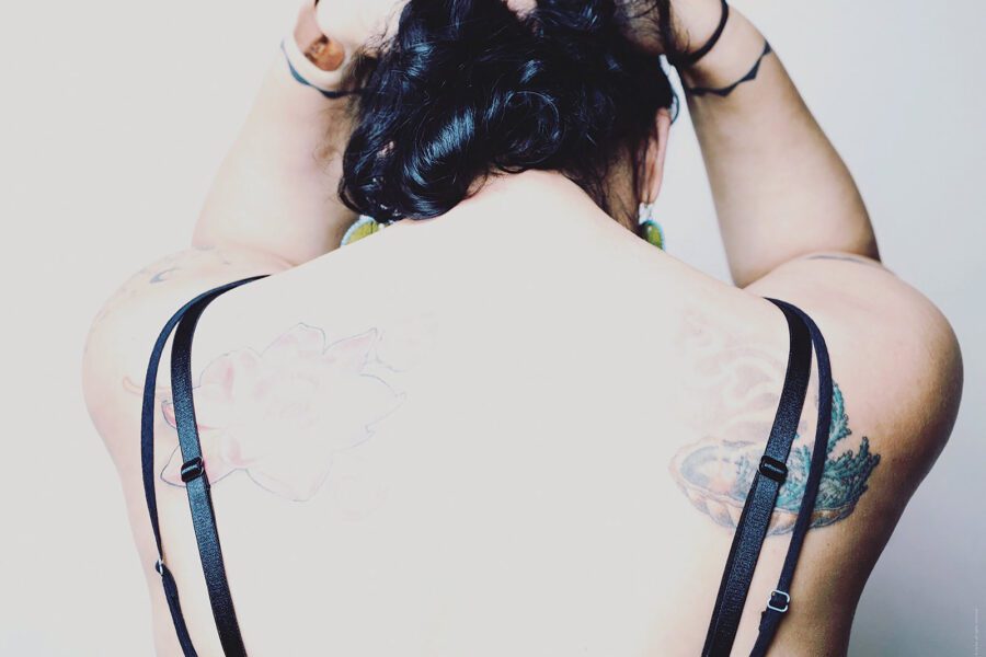 The back of a woman who is facing away from the camera with her hands on top of her head. She has light skin, black hair and tattoos on her back and arms. She has 4 black straps coming from her clothes.