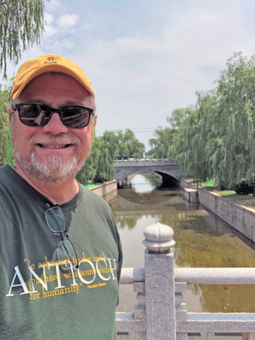 Borrup, clad in a yellow hat, sunglasses, and an Antioch t-shirt, stands on a bridge, looking down a river at another arching stone bridge.