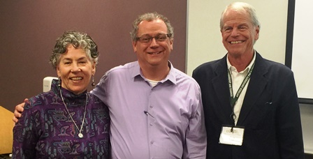 Tom Borrup wears a purple, collared shirt with the top button open, grinning, as he stands between two of his advisors, also smiling.