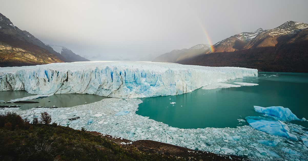 large iceberg in a small inlet, with a rainbow in the distance.
