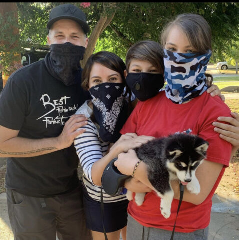 Four people pose for a photo, each wearing facemasks. The one closes to the camera holds a small, cute dog.