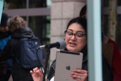 A woman wearing glasses speaks into a large microphone, evidently reading from an ipad she holds in her hand.