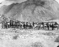 A black-and-white portrait of a group of about 50 people, some on horseback, posing below a mountain range.