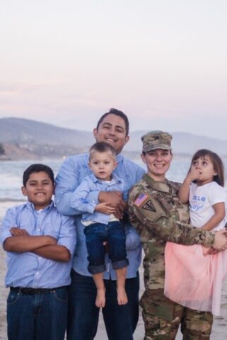 A family portrait with, from left, a boy, maybe ten, arms crossed, a smiling man holding a perhaps four-year-old boy, and a woman in army combat fatigues holding a perhaps six-year-old girl in a pink tutu.