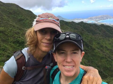 Two women, evidently in the middle of a hike, post for a moutainside photograph, with a sparkling blue bay distantly in the background.