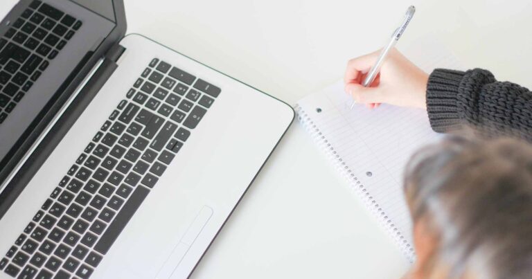Person sitting at a desk in front of a laptop writing in a notebook.