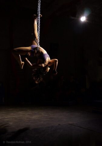 A woman hanging by a chain on a dramatically-lit stage, arches her back in a pose as part of a performance.