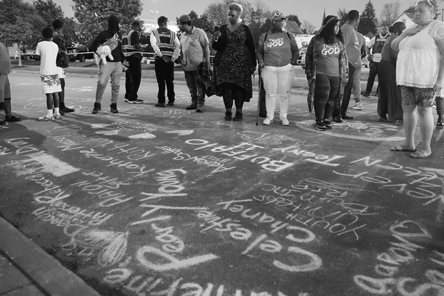 A black and white image of people gathered around sidewalk chalk letters spelling out the names of the Buffalo victims