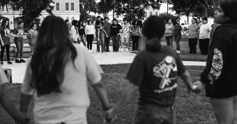 A black and white image of a circle of young and older people in Uvalde, Texas, holding hands after a gun massacre at an elementary school.