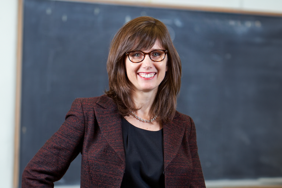 A photograph of Heather Cheney, a white woman wearing glasses, standing in front of a chalk board.