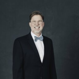 Sean Creighton smiling wearing a bow tie and black suit standing against a dark wall. 