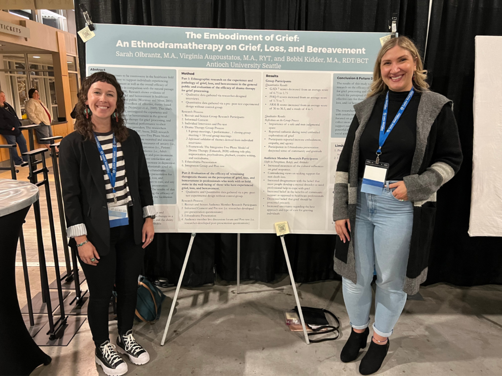 Virg Augoustatos and Sarah Olbrantz standing in front of a presentation board with the title The Embodiment of  Grief An Ethnodramatherapy on Grief, Loss, and Bereavement