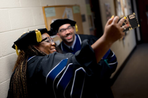 Two people in graduation robes smiling and holding a smartphone, capturing a joyful selfie.