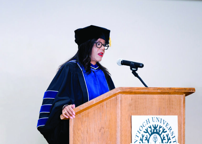 A photograph of Carol Isom-Barnes standing in full regalia at a lectern, giving a graduation speech.