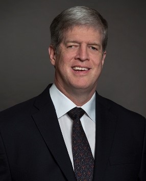 An photo of Kirk Adams. He is smiling and wearing a black jacket, white shirt, and dark tie. The background is a dark gray.