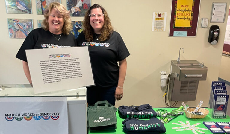 Antioch University staff hosting a table for voting. Table has a green cloth, with swag, such as T-shirts on the table.