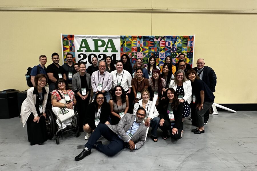 The PsyD group that went to the APA conference. A diverse group of individuals smiling and posing together in front of a colorful banner.