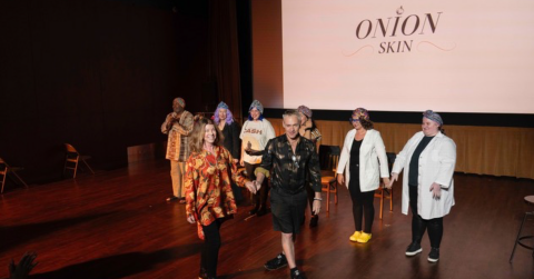 A diverse group of individuals stands on a stage in front of a large projector screen, engaging with the audience. The screen says Onion Skin