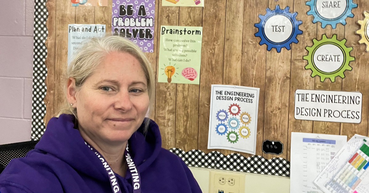 A woman wearing a purple shirt sits in front of a bulletin board, engaged in thought or discussion.