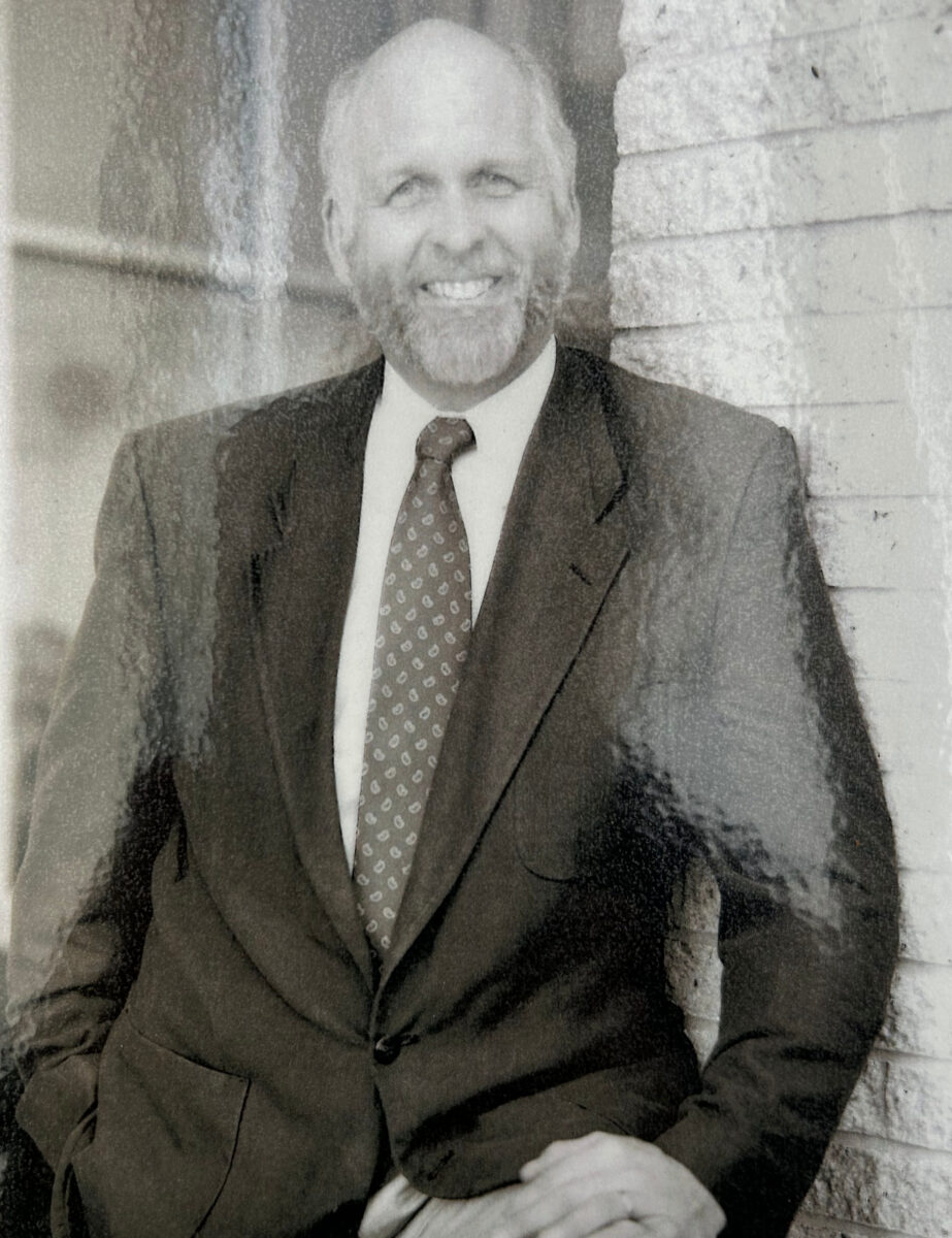 A black and white photo of Jim Craiglow. He is standing next to a brick wall and wearing a suit. 