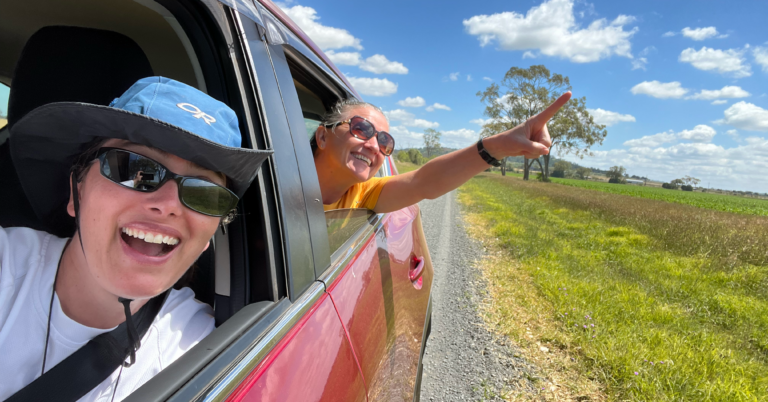 A photograph of Liz Foote (in front) and her friend Sharyn spotting a koala in the wild, while driving down the road. Sharyn points out of the back seat with great enthusiasm.