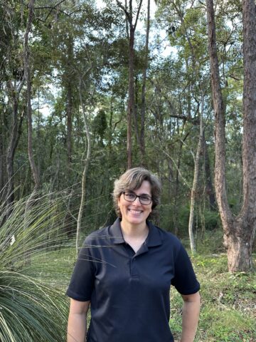 A photograph of Liz Foote standing out in the forest, wearing a black polo shirt and eyeglasses.