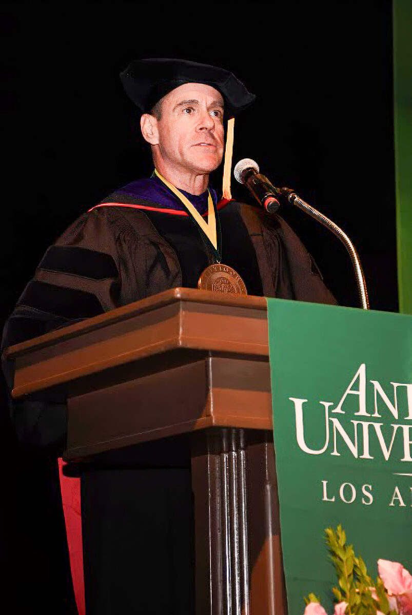 Chancellor Bill Groves, a white middle-aged male, at a podium during a commencement ceremony. He is wearing regalia. 