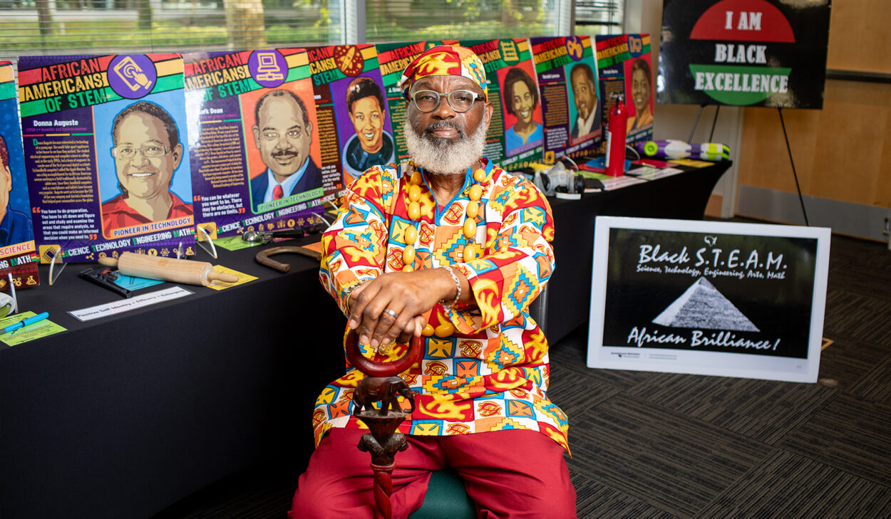 Delbert Richardson, an african american male, sitting in front of posters for "African Americans in Stem" exhibt. He is wearing brightly colored attire with an ornate cane