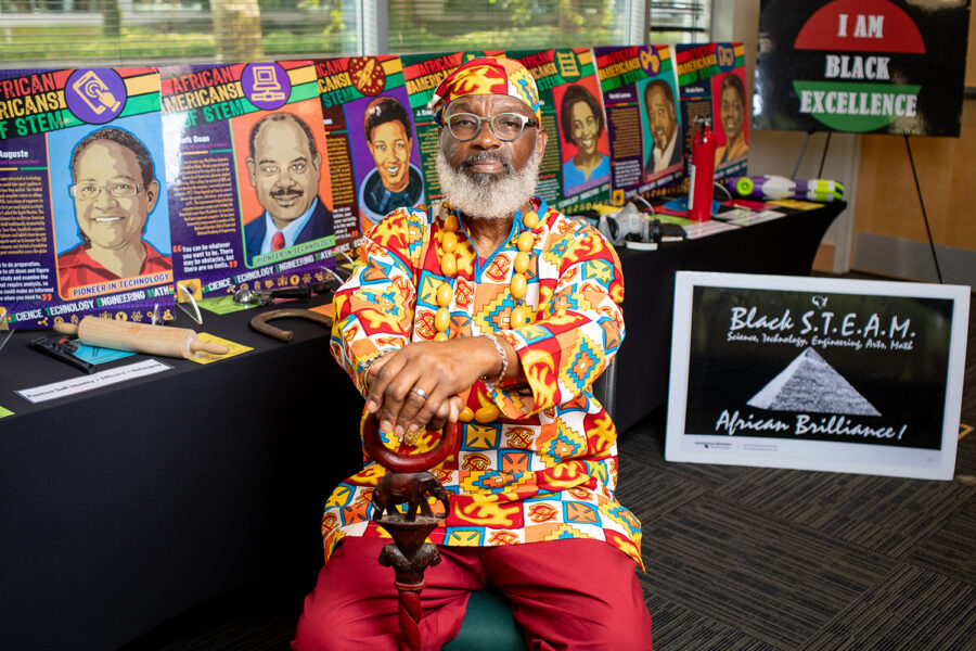 Delbert Richardson, an african american male, sitting in front of posters for "African Americans in Stem" exhibt. He is wearing brightly colored attire with an ornate cane