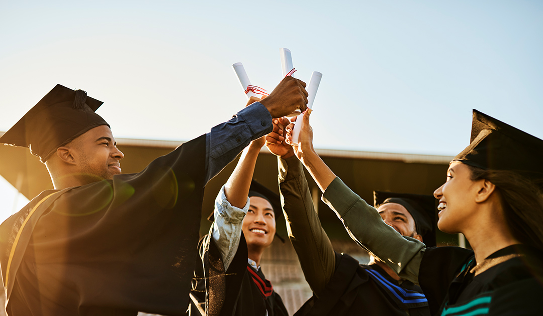 A group of 4 graduates with their hands and diplomas up and meeting in the middle of the circle.
