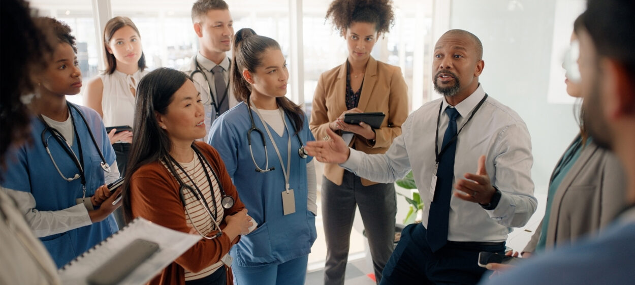 A group of diverse people in a medical styled office setting, listening to one person speaking.