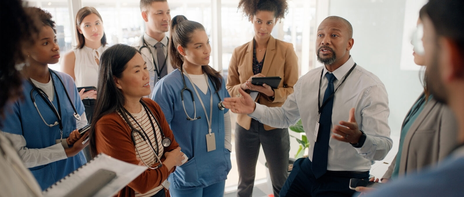 A group of diverse people in a medical styled office setting, listening to one person speaking.