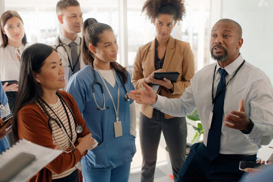 A group of diverse people in a medical styled office setting, listening to one person speaking.