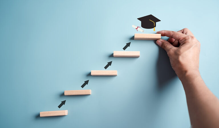 A hand reaches for a graduation cap resting on a wooden stair, above several other wooden stairs, symbolizing achievement and aspiration.