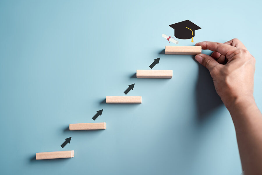 A hand reaches for a graduation cap resting on a wooden stair, above several other wooden stairs, symbolizing achievement and aspiration.