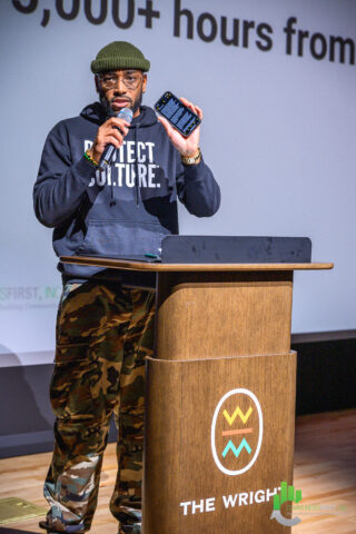 A photo of a man, Lance Wheeler, the Vice President of Learning and Engagement at Charles H. Wright Museum of African American History in Detroit, standing at a lectern, giving a speech. He's holding a microphone and a cell phone. The lectern says "THE WRIGHT" and has a logo for the museum.