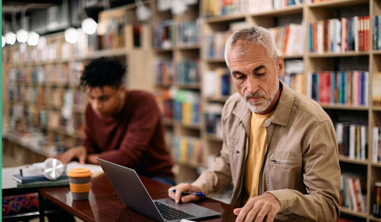 A man focused on a laptop while seated at a table in a quiet library setting.