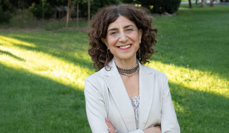 A horizontal image of a woman in a white blazer standing in front of green grass with dappled light on a out of focus background