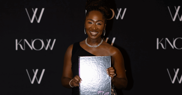 Stephanie Helms Pickett, wearing a black one-shoulder dress and silver jewelry, smiles while holding a reflective silver book in front of a black backdrop with repeating “KNOW” and “W” logos.