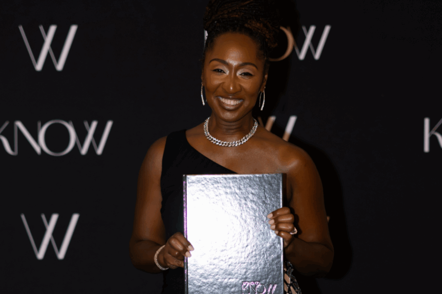 Stephanie Helms Pickett, wearing a black one-shoulder dress and silver jewelry, smiles while holding a reflective silver book in front of a black backdrop with repeating “KNOW” and “W” logos.