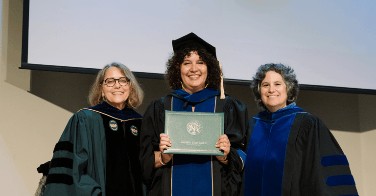 A photograph of Alicia Wargo standing on stage in full PhD regalia, flanked by two of her professors.