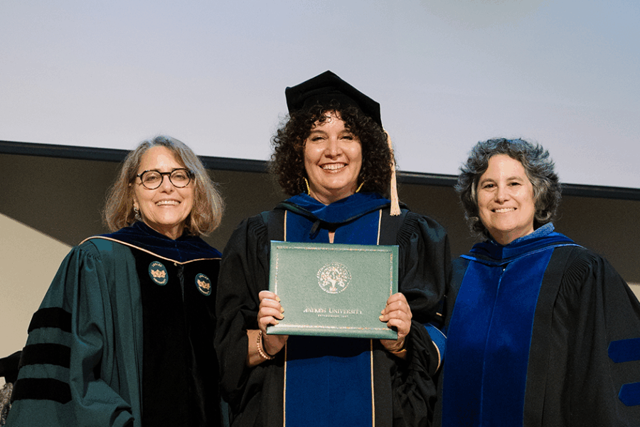 A photograph of Alicia Wargo standing on stage in full PhD regalia, flanked by two of her professors.