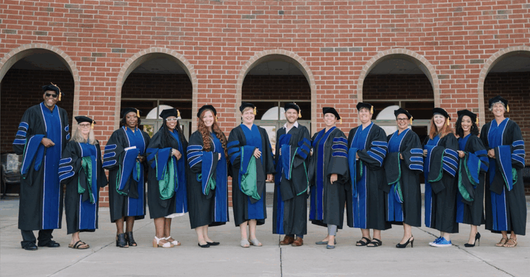 A photo of about a dozen Antioch graduates in their PhD regalia standing in front of a brick building with archways.