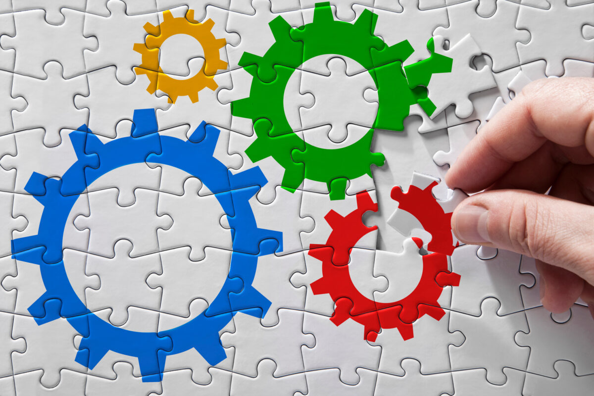 A white puzzle with gears, blue, orange, green, and red. A hand is completing the final pieces. `