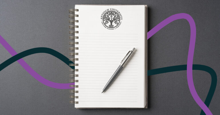 An open spiral notebook with lined pages and a silver pen resting on it. At the top of the page is the Antioch University logo with the words “Uniting Passion & Purpose.” The notebook is set on a gray background with purple and teal wave-like lines flowing across it.